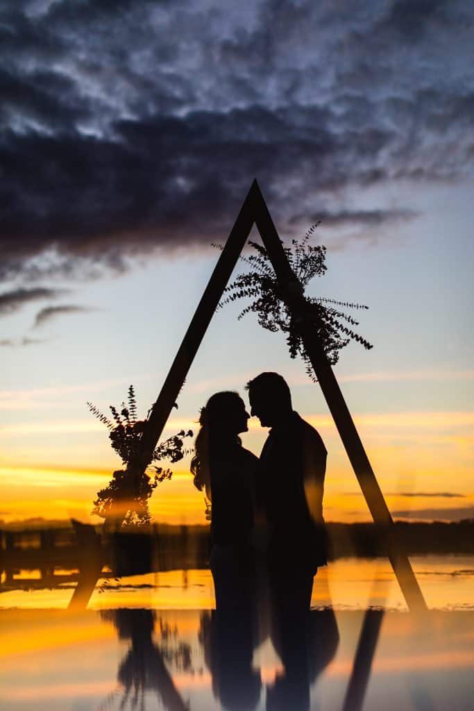 man and woman embracing at the edge of the water during a beautiful sunset with a reflection of an alter, Lavender on the Lake, Orlando, FL