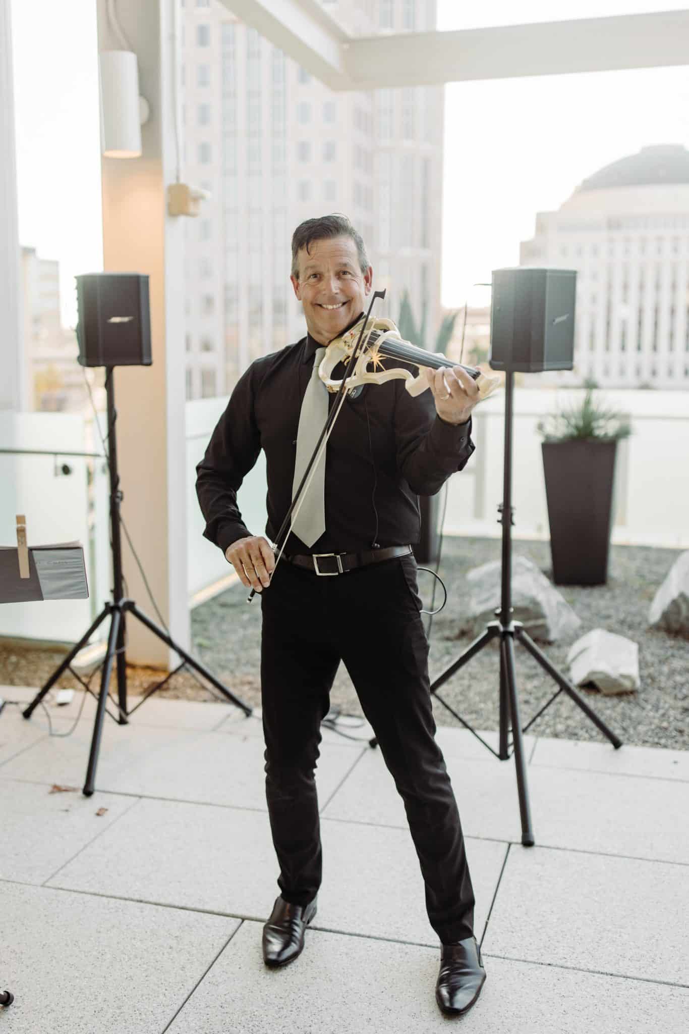 man wearing all black stands in front of speaker smiling playing violin