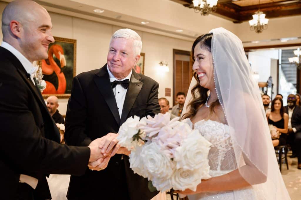 dad passes bride over to groom on wedding day during the ceremony while she holds white bouquet of flowers