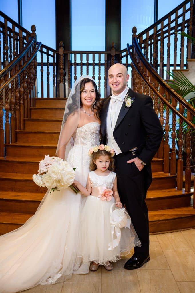 bride and groom pose with flower girl in front of staircase on wedding day