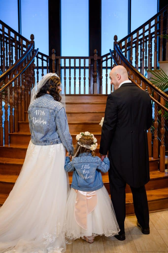 bride and flower girl stand with groom wearing matching jean jackets facing away from the camera