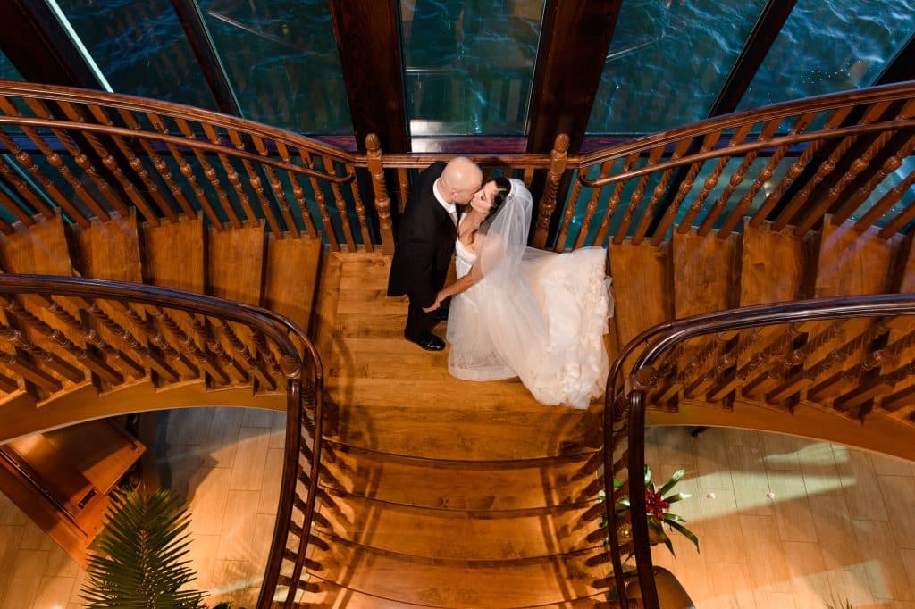 bride and groom kiss in the middle of wooden staircase with water in the background of windows