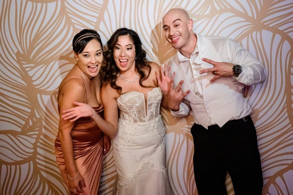 bride groom and one other woman stand in front of tropical gold floral print backdrop in photobooth