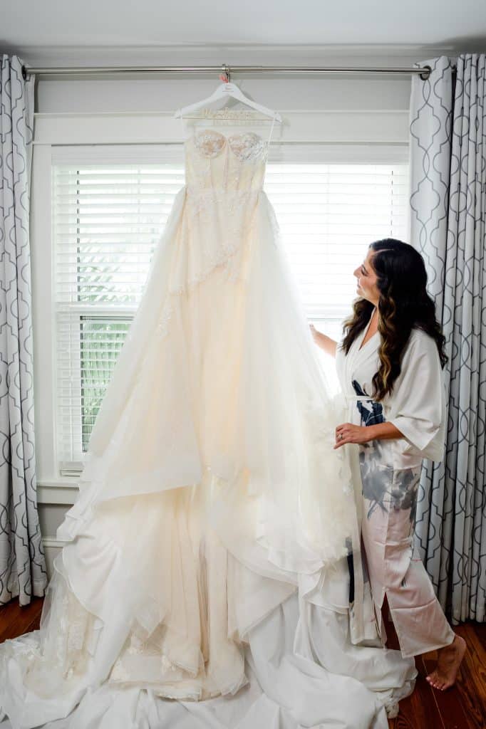 bride stands in front of window looking at wedding dress hanging up