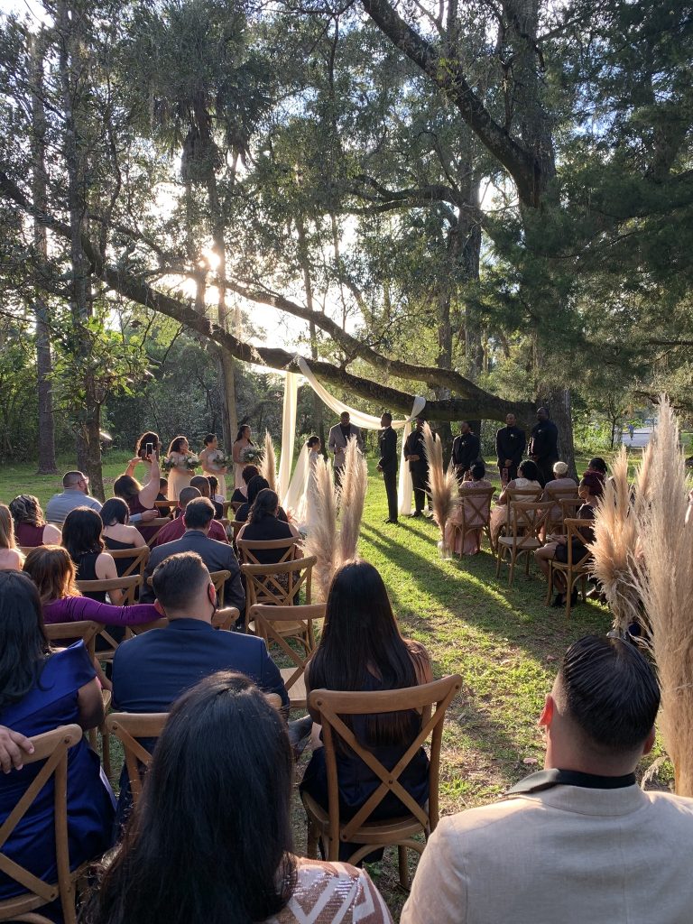 outdoor wedding ceremony nearing sunset, in the woods, under trees with sunlight coming through, wedding guests in wooden chairs, Orlando, FL