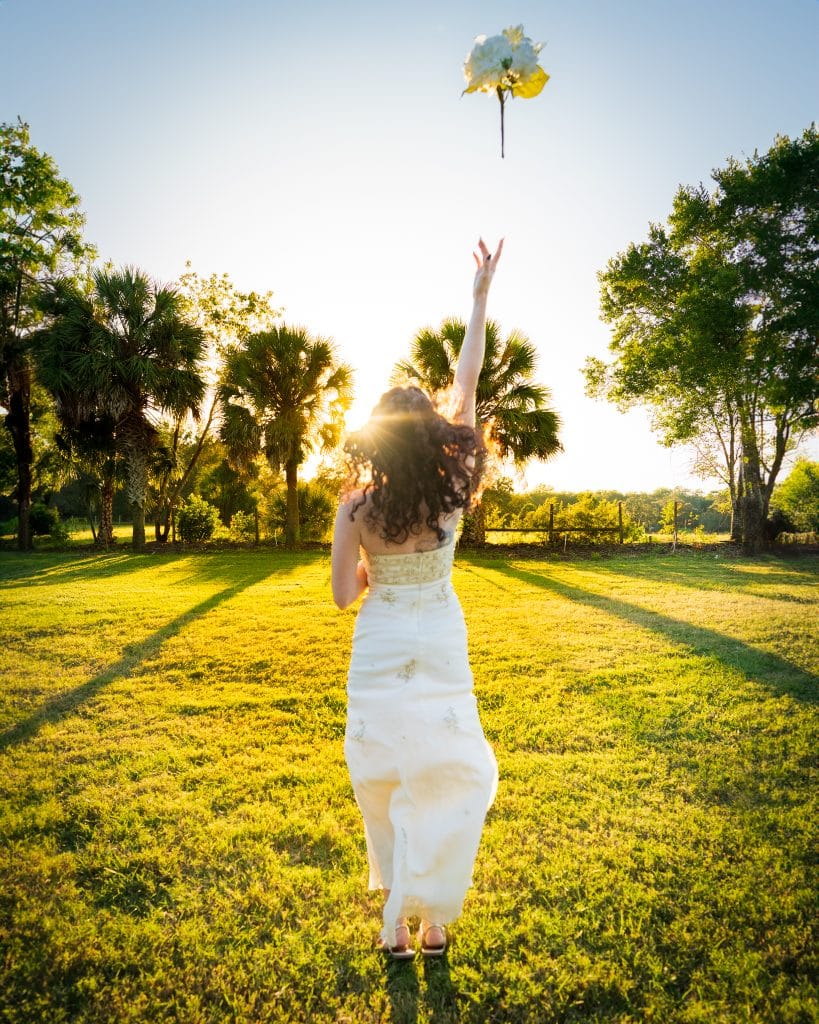 bride throwing her white flower bouquet into the air in a yard with trees surrounding her, orlando, FL