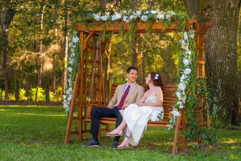 couple sitting on wooden bench swing, outdoors, under a tree, draped in greenery and white flowers, orlando, FL