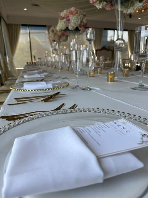 Place setting on table, white chargers, white napkins, menu cards, tall flower centerpieces, glass stemware, gold flatware, One Night Only, Orlando, FL