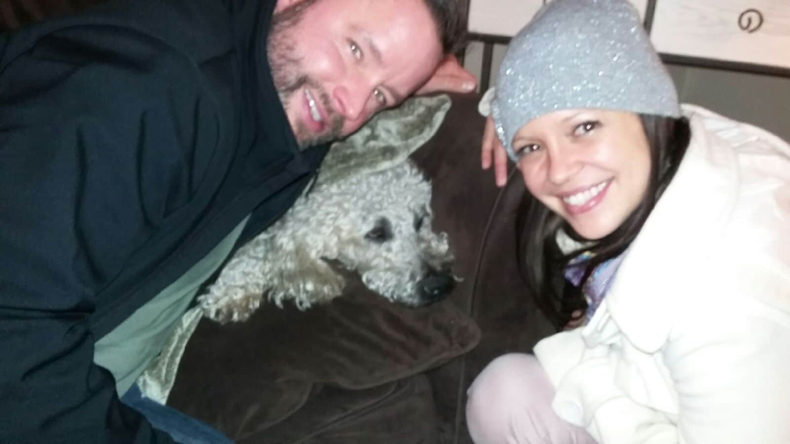 man and woman lean over couch with dog laying on it smiling for picture