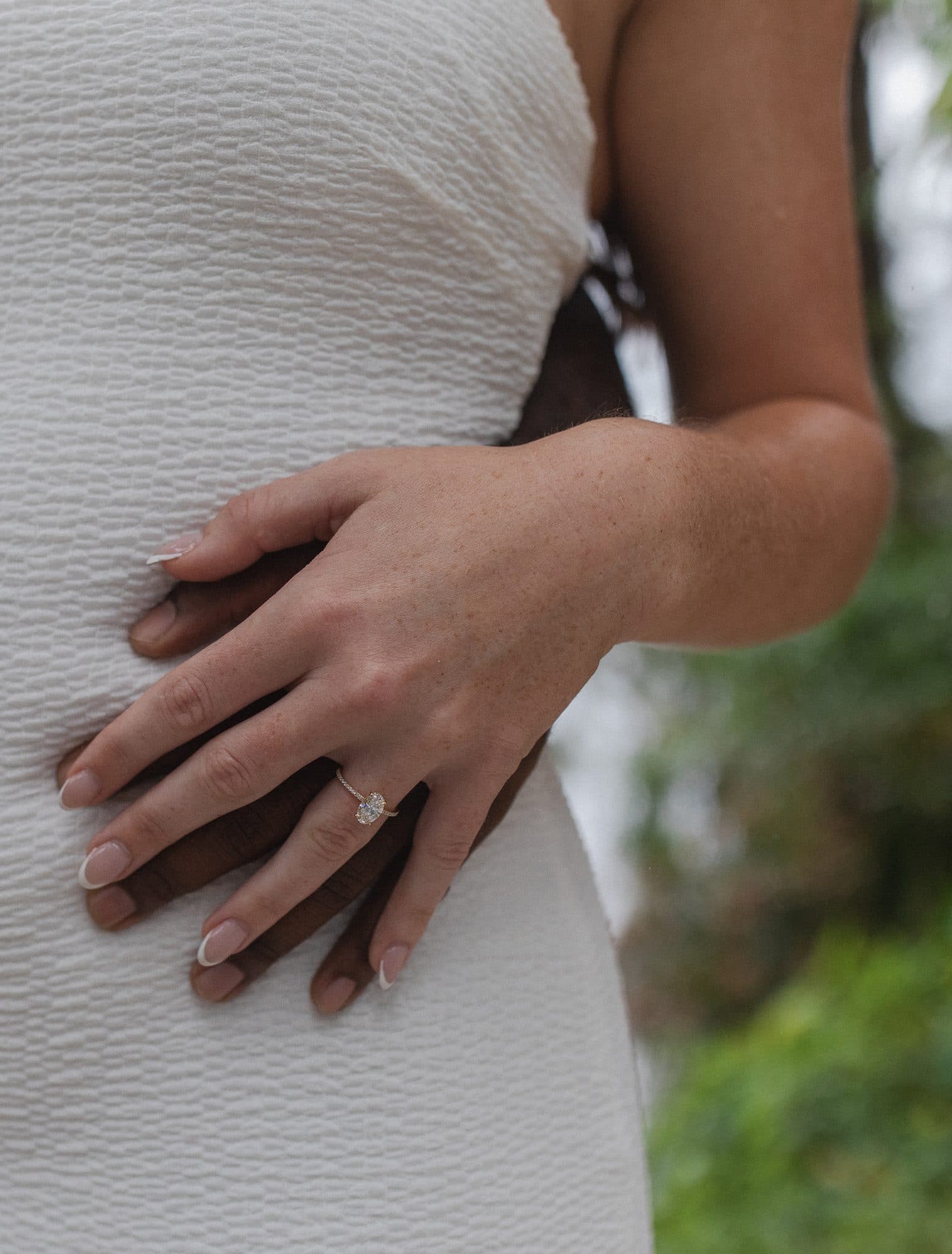 close up of mans hand on womans waist with her hand on top showing oval engagement ring