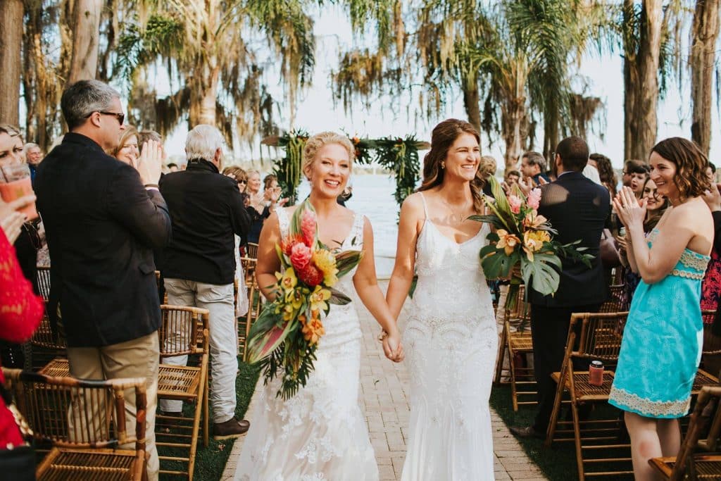 two brides walking down the aisle, outdoors under palm trees, guests applauding, paradise cove, central FL