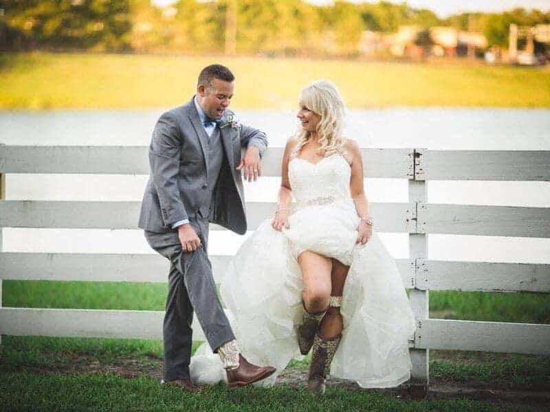 wedding couple in their wedding attire, standing against a white fence on the ranch, bride in her wedding gown with cowboy boots, groom in a grey suit, P.S. I Love You Weddings & Events, Orlando, FL