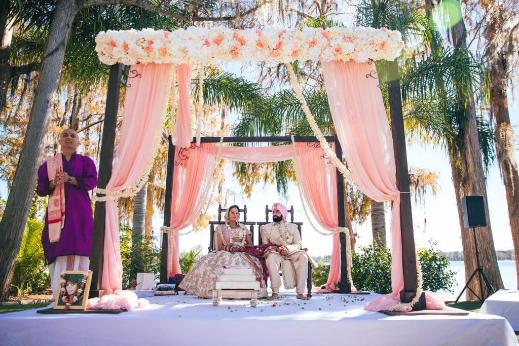 bride and groom sitting under the altar dressed with pink material and white and pink flowers, outdoors, palm trees, beach, central fl