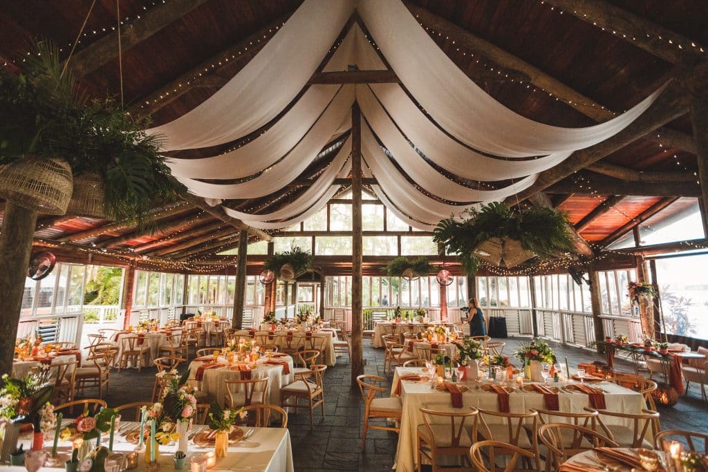 indoor venue, large floor length windows, white tapestry hanging from the ceiling, white tablecloths with maroon runners, gold chairs, Central FL