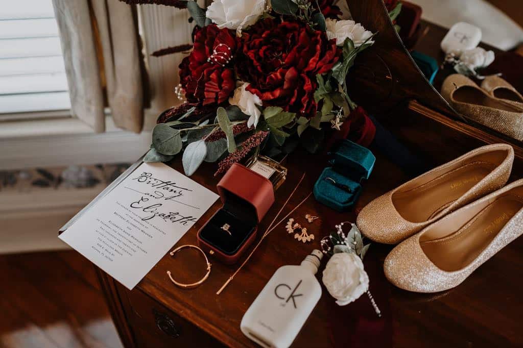 Flatlay of wedding shoes, perfume, invitation, bouquet of red and white flowers, wedding rings, jewelry and corsage, Orlando, FL