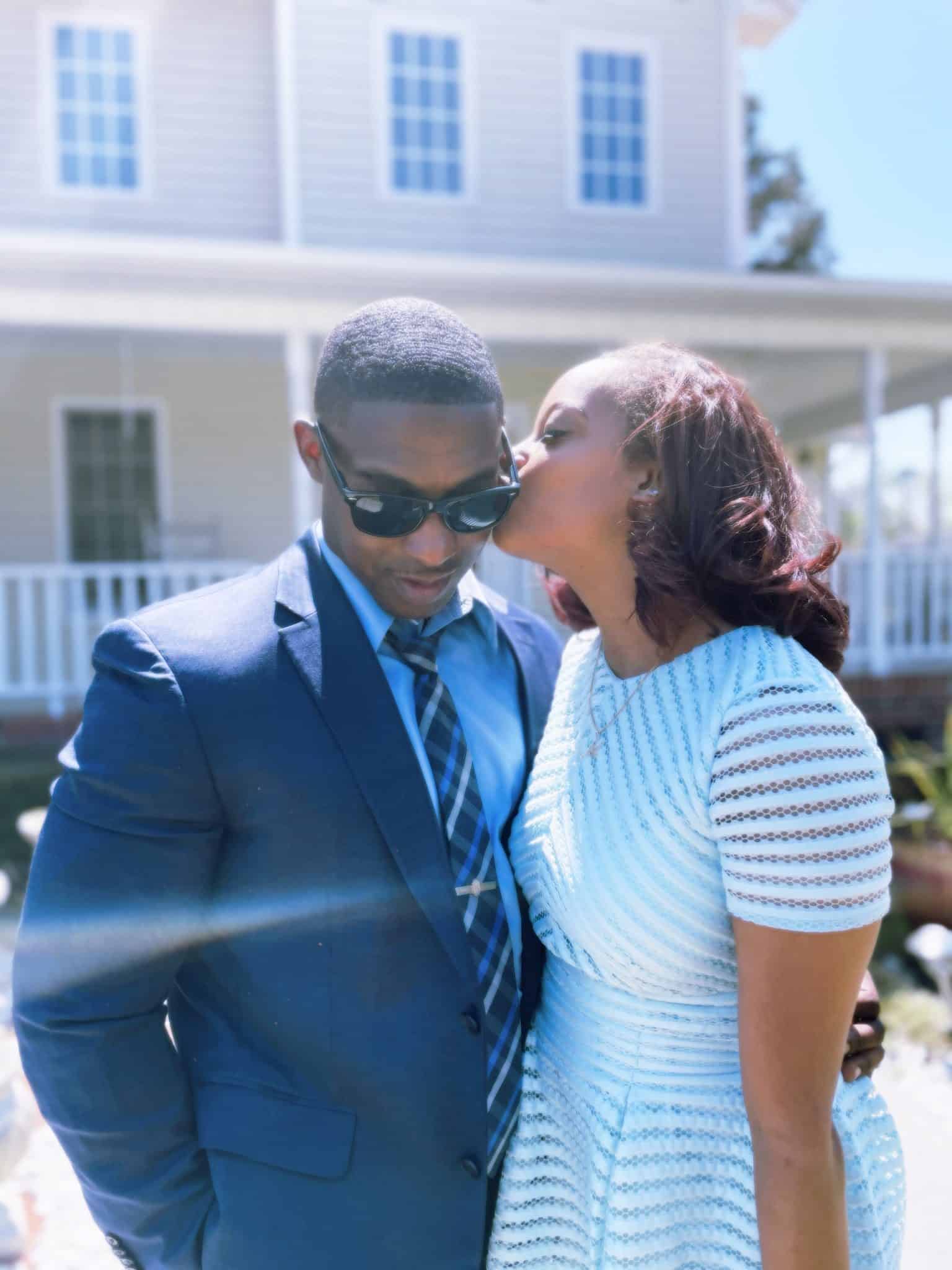 man and woman stand together while woman kisses mans cheese in front of home