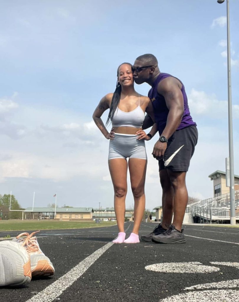 man and woman stand posing on running track outside while man kisses cheek of woman