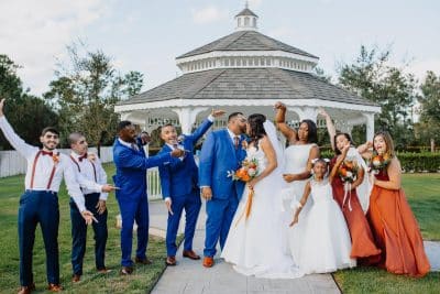 Wedding party celebrating on the lawn at The Center at Deltona in Central Florida, with groomsmen in royal blue suits, bridesmaids in rust and terracotta gowns, and the bride and groom sharing a kiss in front of a white gazebo