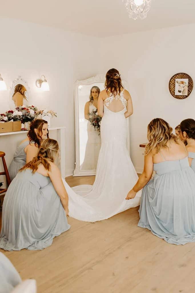 Bride looking in a full-length mirror surrounded by her bridesmaids. They are helping fan her dress out