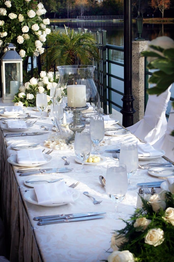 tablescape, large white candle in a clear globe vase on the center, white roses on each end of the table, white tablecloth, outdoors on the patio, Orlando, FL