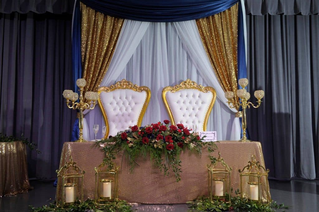 head table set with a tan tablecloth, red flower arrangement, white candles on the floor, white cushion backed chairs with gold trim, blue, gold and white curtains, The Center at Deltona, Orlando, FL