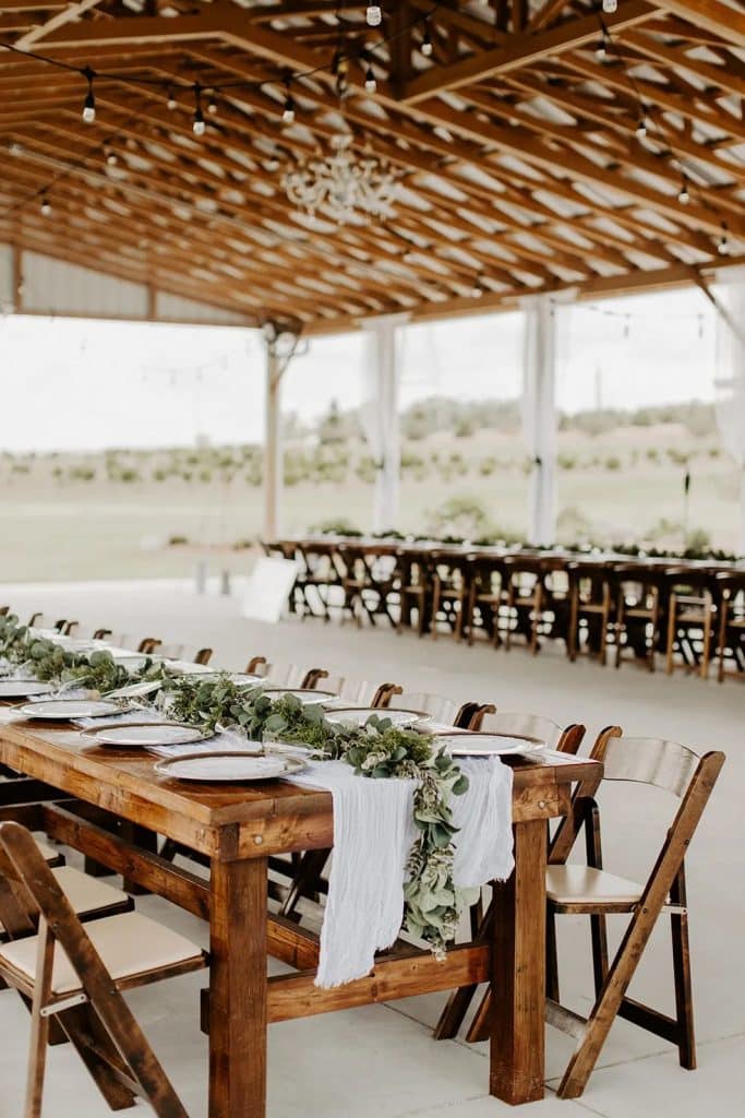 Reception tables with white linens and green garland on wooden tables surrounded by wooden chairs