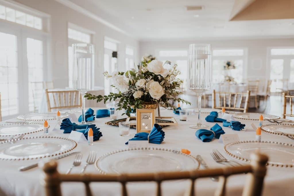 close up of a table set for a reception with a view that includes gold chairs, round table, white tablecloths, blue napkins, white flower centerpieces, clear chargers, Orlando, FL