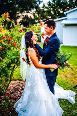 Bride and groom share a romantic moment surrounded by lush greenery and red florals at The Barn at Southern Oaks, a rustic barn wedding venue in Mount Dora, Central Florida