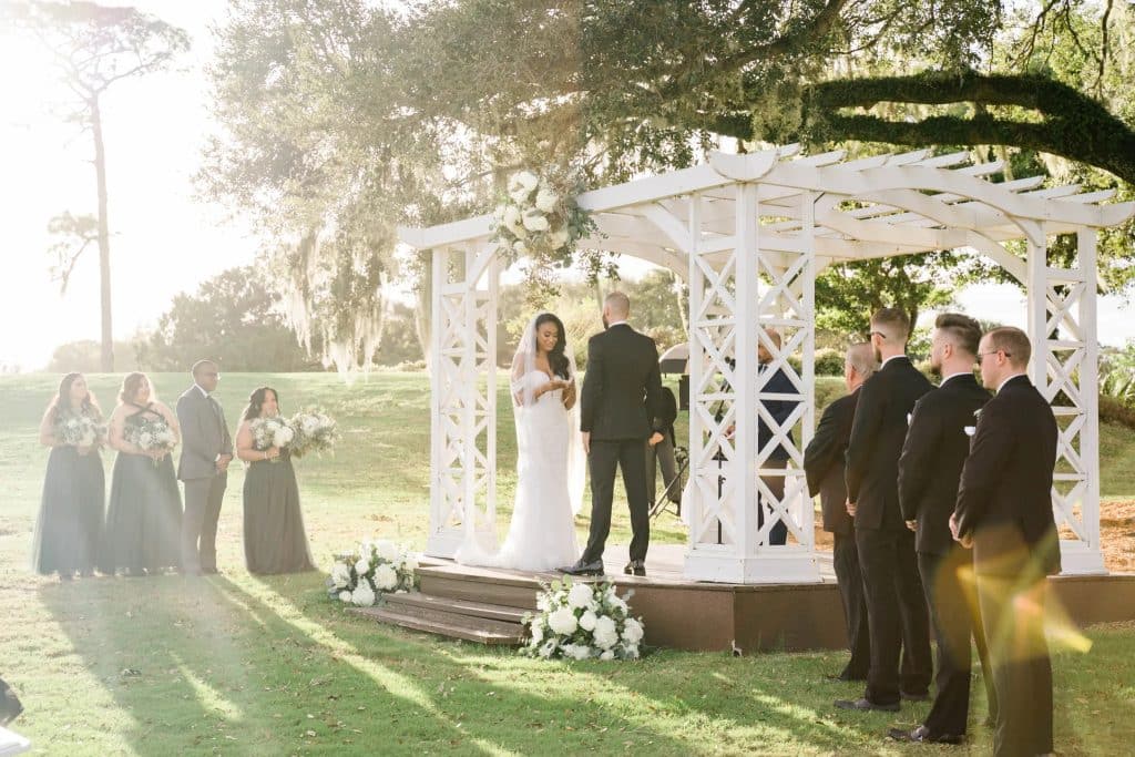 white wooden pergola, outside, wedding couple on the elevated platform, wedding party standing on either side, trees surrounding the area, white flowers, at the bottom of the platform, Tuscawilla Country Club, Orlando, FL