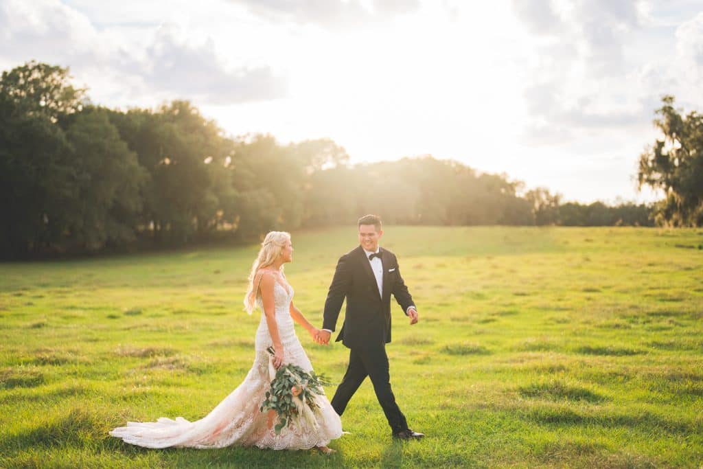 bride and groom walking hand in hand in a green field, clear sky, sunshining, green trees, Central FL