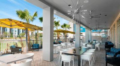 Outdoor covered patio dining area at Hyatt House Orlando Airport featuring white tables and chairs, yellow market umbrellas, modern sputnik chandelier, and tropical palm trees, an affordable casual wedding venue in Orlando Central Florida