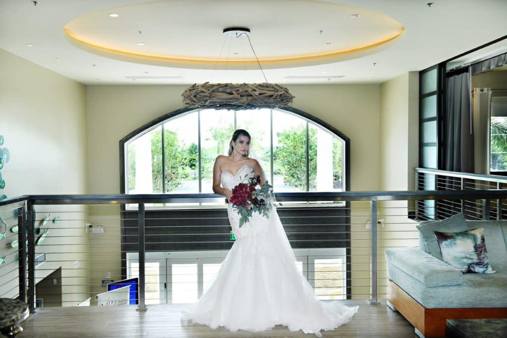 Bride standing on the balcony at the reception, holding her bouquet, large window in the background, silver railings, facing the room, Rentyl Resorts, Orlando, FL