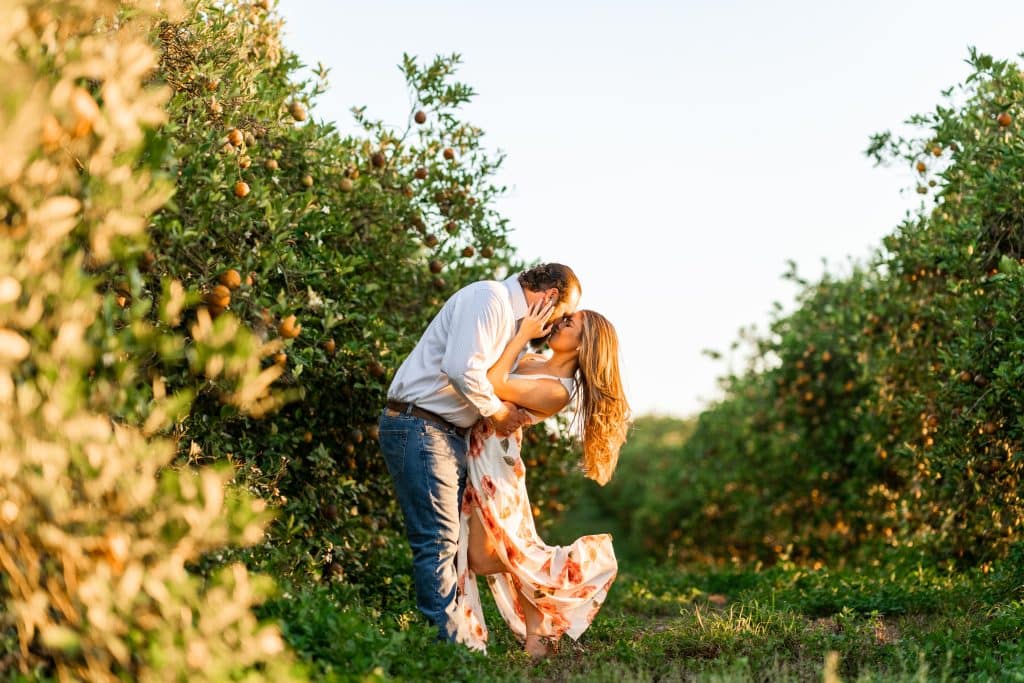 man and woman in an orchard, embracing and kissing, woman in a floral dress, man in dark pants and a white shirt, Polk Bros Photo.Video.DJ, Central FL