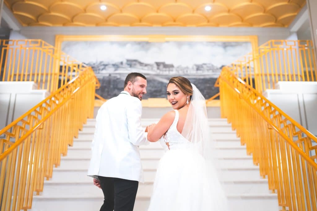 bride and groom ascending a white staircase with gold railings, looking back at the camera, Polk Bros Photo.Video.DJ, Central FL