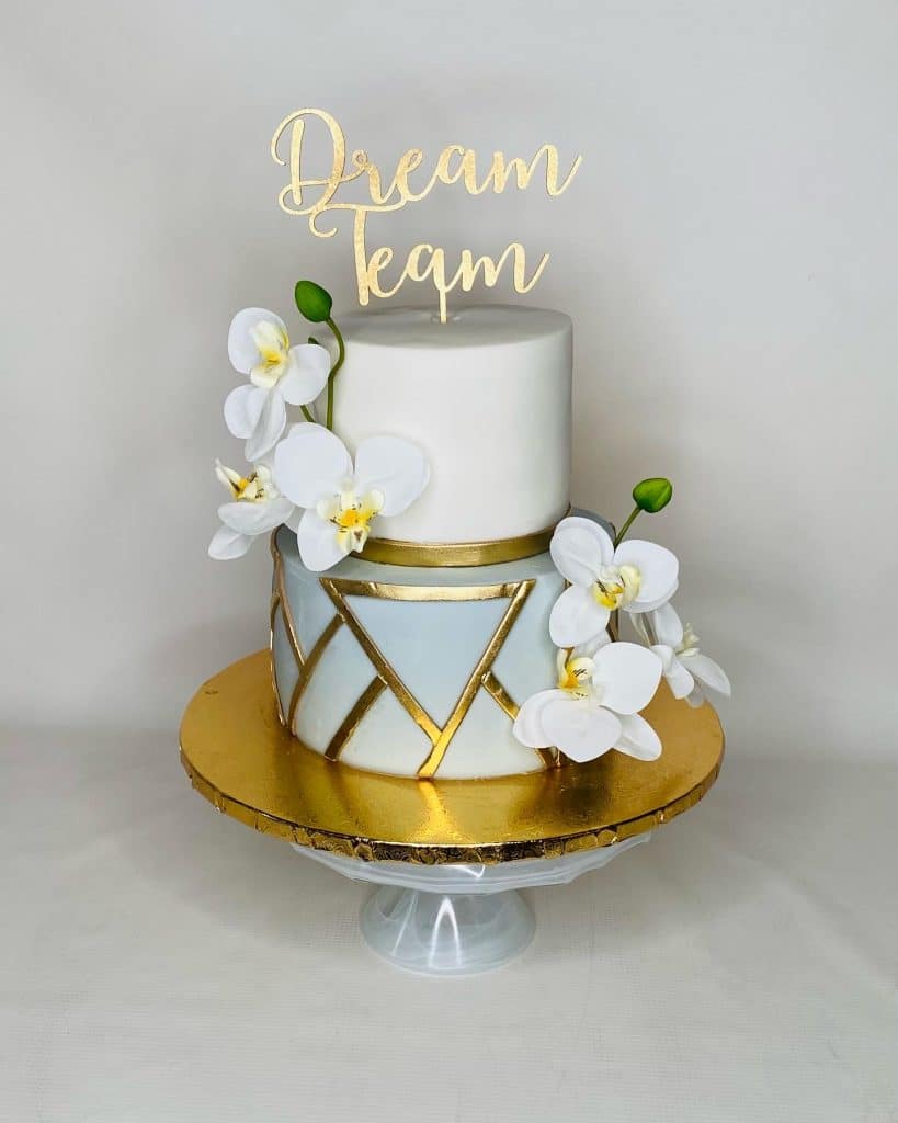 dream team cake, top layer is white, bottom layer is light blue, white flowers adorning the sides, gold cake stand with gold embellishments on the layers, sugar divas bakery, orlando, fl