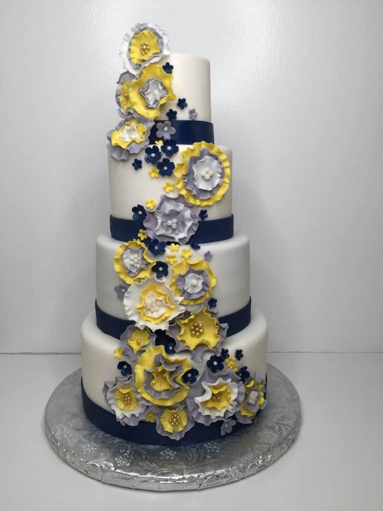 Four layer cake, all white fondant with blue ribbons along the bottom of each layer, yellow and blue flowers covering most of the front fo the cake, silver plate, orlando, FL