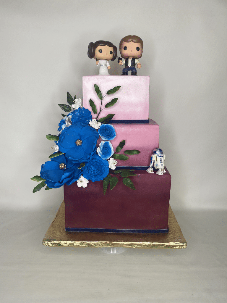 Three layers, square shaped, large blue flowers on the left corner, funky pops on top layer for the bride and groom, R2D2 figurine on the bottom layer, light pink, medium pink and maroon colors for the layers, Sugar Divas Cakery, Orlando, FL