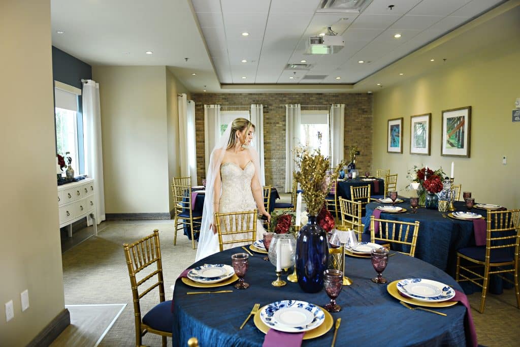 bride wearing her wedding dress, in a small, intimate setting, with round tables, dark blue tablecloths, gold chargers, white and blue plates, gold chairs, red flower centerpieces, Orlando, FL