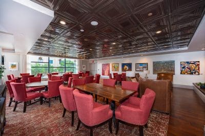 paintings on the far wall, lounge area with red chairs and tables, partial wood floors, partial rug under the tables, large windows, Orlando, FL