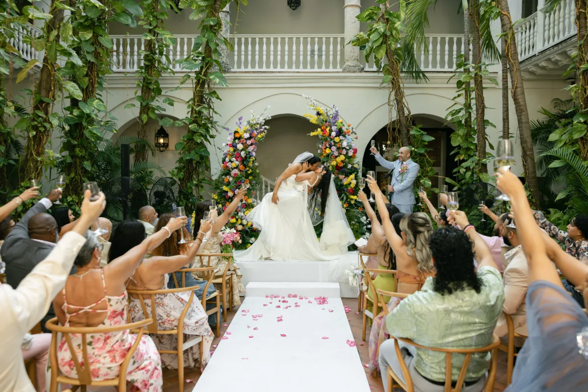 Orlando wedding photographer Nexum Photography capturing joyful same-sex wedding ceremony with two brides kissing under a colorful floral arch as guests cheer and toast in a tropical courtyard venue