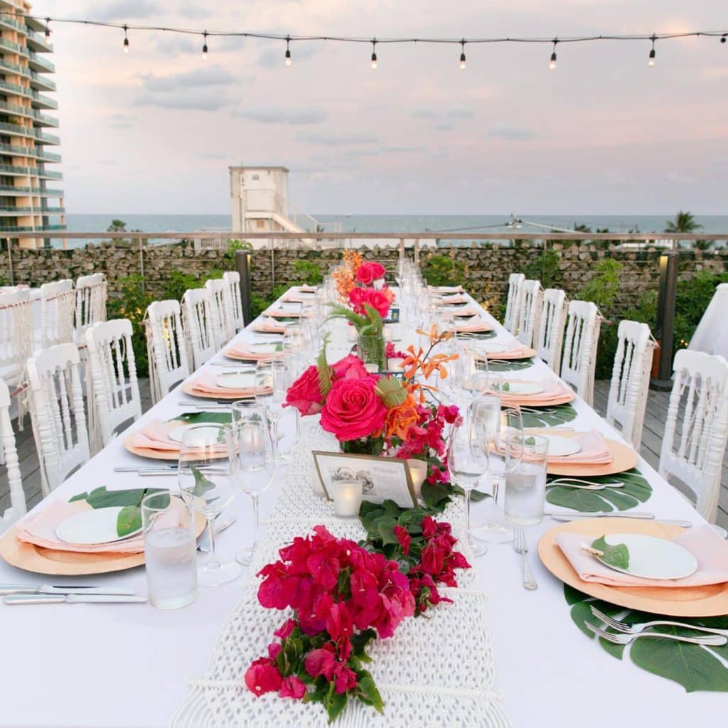 rooftop area, string lights hanging above, long table, red flowers as centerpieces down the middle, white chairs, white tablecloth, green leaf placemats, light pink plates and bowls, Naomi Zora Events, Orlando, FL