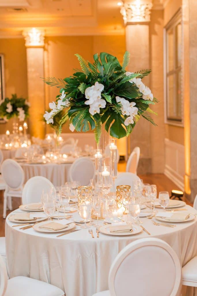 close up of a round table set for a reception, tall centerpiece of white flowers with greenery, white tablecloths, white chairs, white china and candles, Orlando, FL