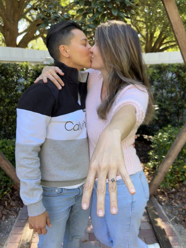 couple kisses outside while one holds hand out with engagement ring on