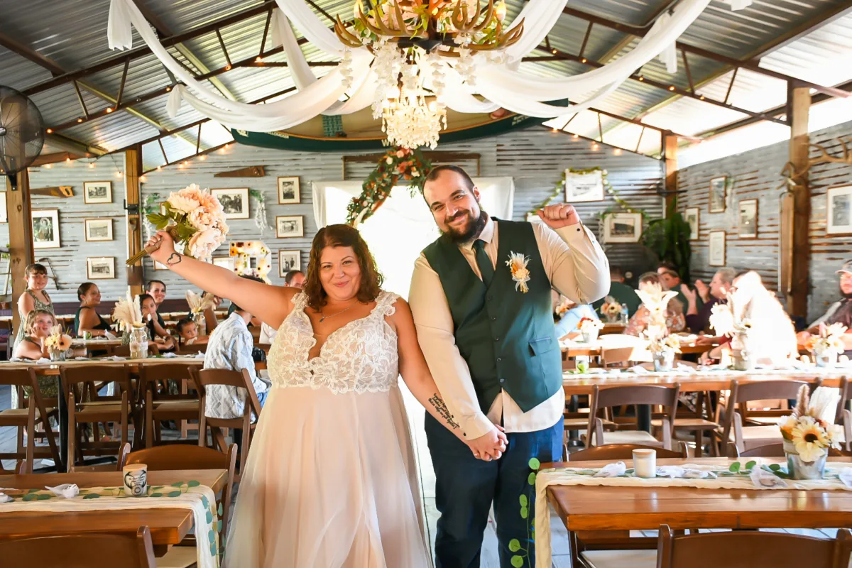Bride and groom celebrating joyfully at their rustic barn wedding reception at S&S Ranch Wedding and Entertainment Venue in Central Florida, bride holding bouquet overhead in a lace wedding gown as groom in green vest raises fist in celebration surrounded by seated guests at wood farm tables decorated with sunflowers and greenery