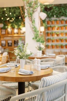 Elegant outdoor dining setup at Summer House on the Lake in Orlando Florida featuring round wooden tables with blue striped wicker chairs, fresh wildflower centerpieces, and a lush living wall of terracotta pots as a backdrop for this boho wedding venue