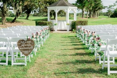 Outdoor wedding ceremony setup at Falcon's Fire Golf Club in Kissimmee Florida featuring white folding chairs lining a grass aisle adorned with pink and mauve floral arrangements leading to a white gazebo surrounded by lush golf course greenery