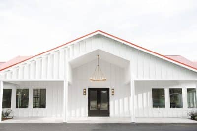 The Colonial at Sunset wedding venue exterior featuring white board and batten siding with a red metal roof and elegant chandelier in the covered entrance, a modern farmhouse event space in Central Florida and Tampa
