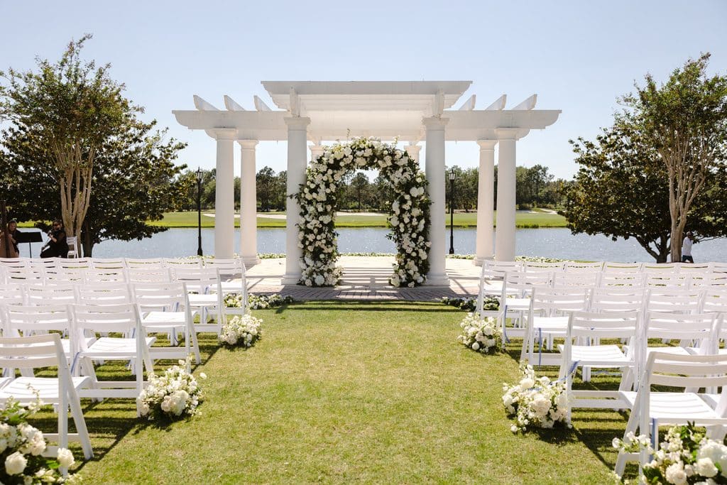 wedding ceremony set up, outdoors, white chairs in rows with a center aisle, altar at the front, adorned with all white flowers, green background, white pergola, water beyond the altar, Orlando, FL