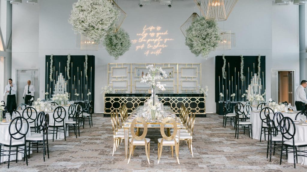 reception set up, round tables on the left and right with white tablecloths and black chairs, down the center is a long rectangle table with gold chairs and white tablecloth, large topiaries hanging from the ceiling, Orlando, FL