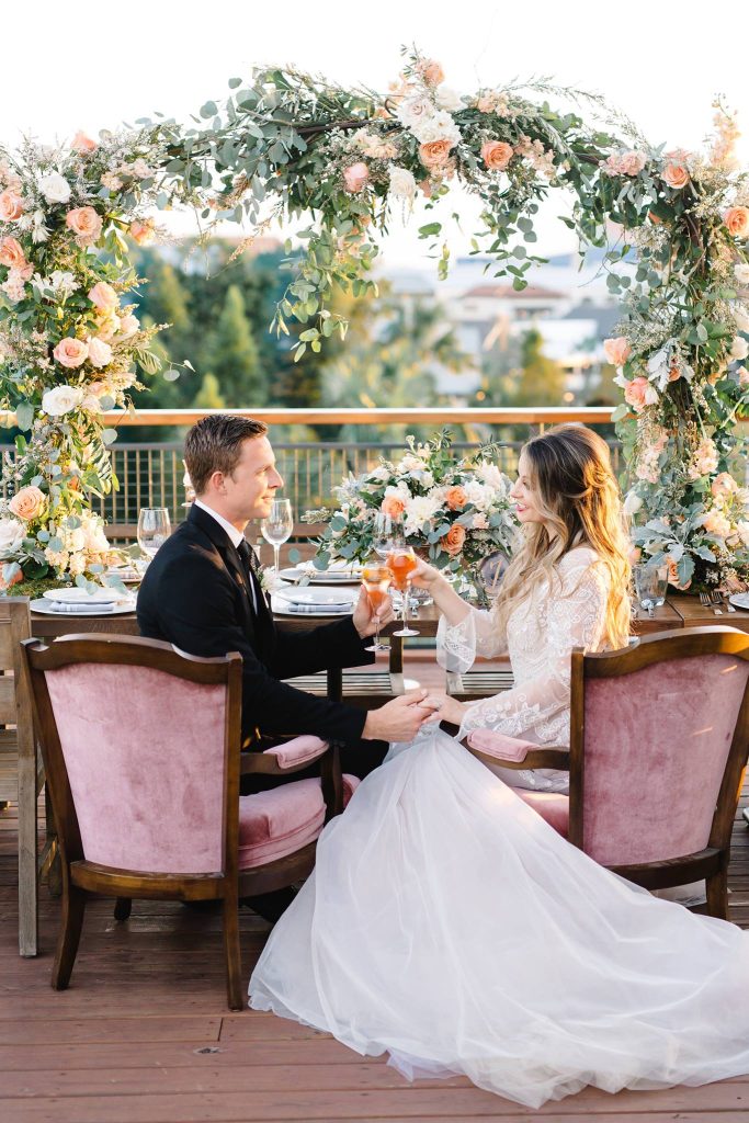 bride and groom sitting at their head table during the reception, pink chairs, floral arrangement arching above, Orlando, FL